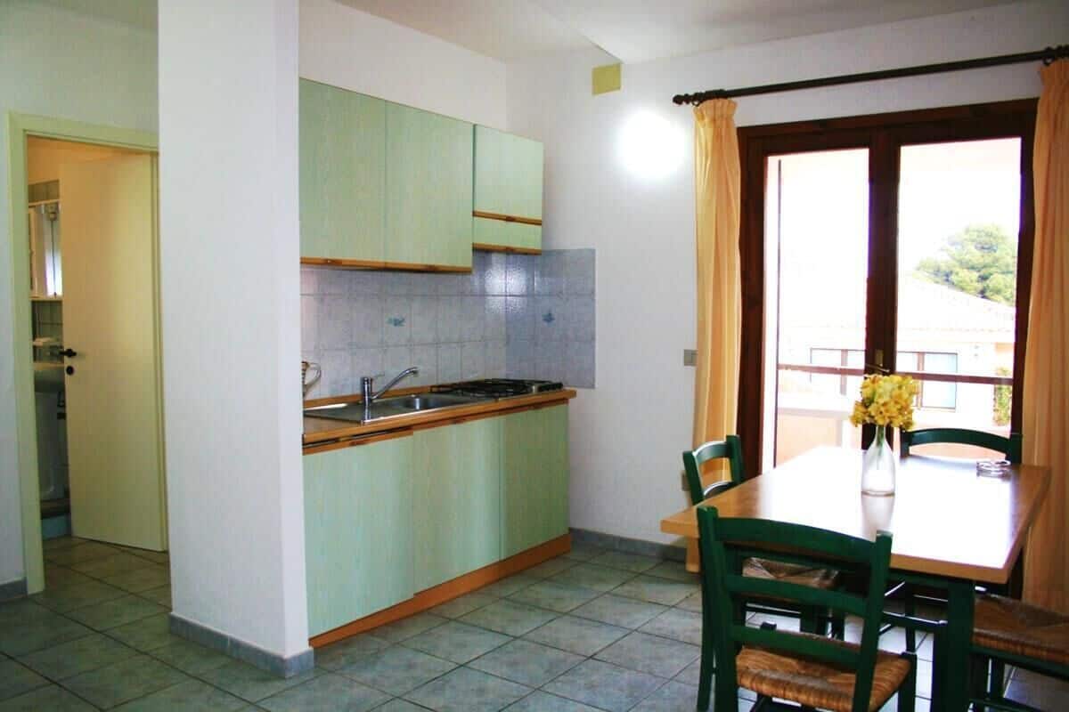 Una cocina pequeña con gabinetes verdes, un lavamanos y una estufa. Hay una mesa de comedor con sillas y un florero con flores.
