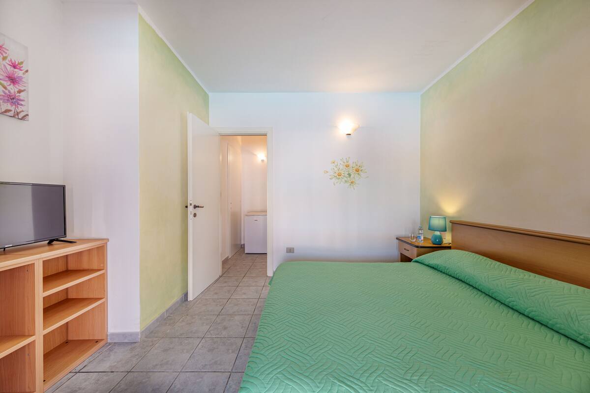 Un dormitorio con una colcha verde, un televisor sobre una estantería de madera, una mesita de noche con una lámpara, y una puerta que da a un baño.