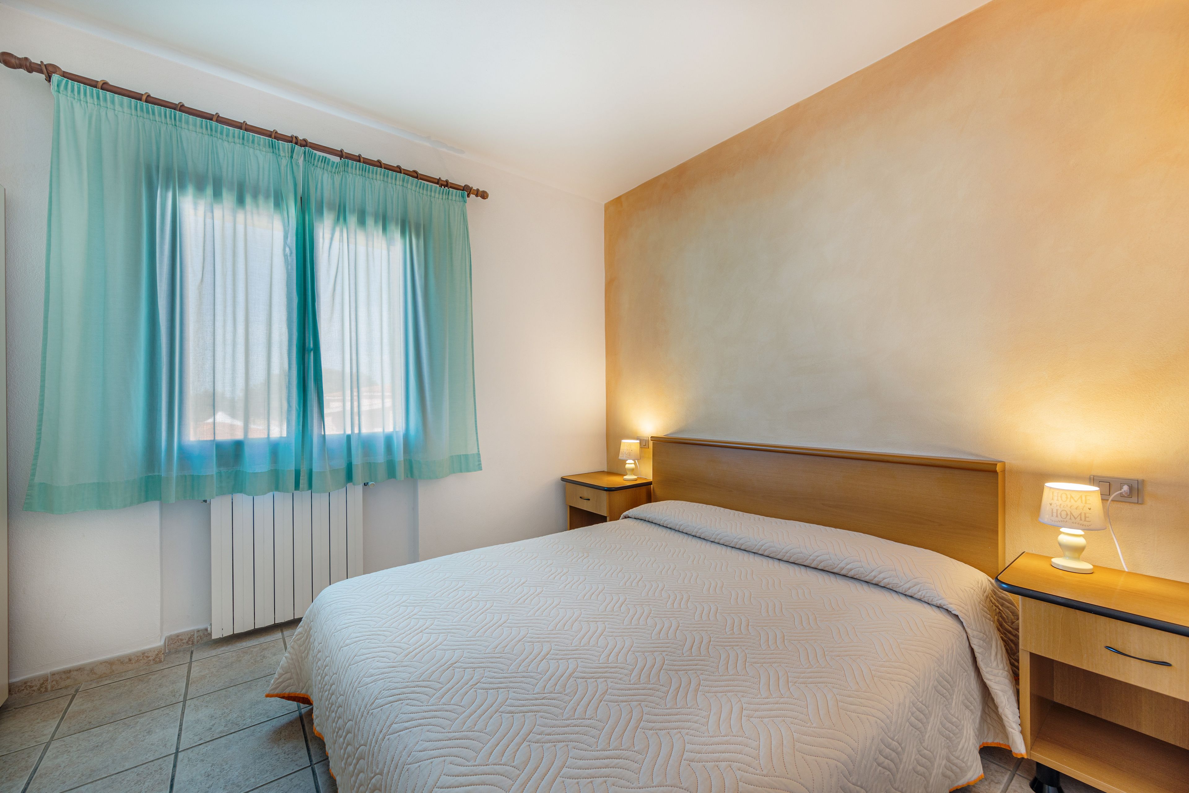 A hotel room with a large bed, a nightstand with a lamp, a window with curtains, and a radiator.