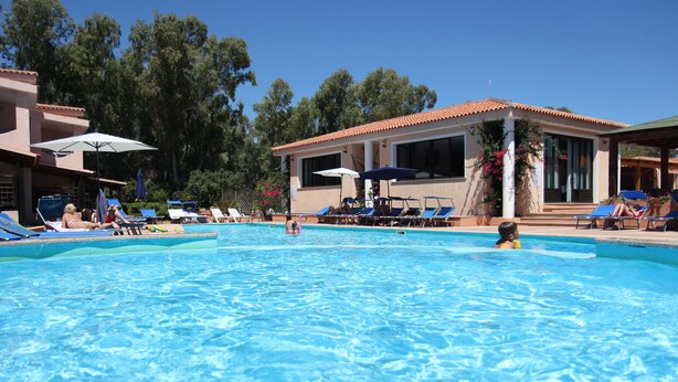 Un espace piscine doté de chaises longues et de parasols, entouré d’immeubles et d’arbres.