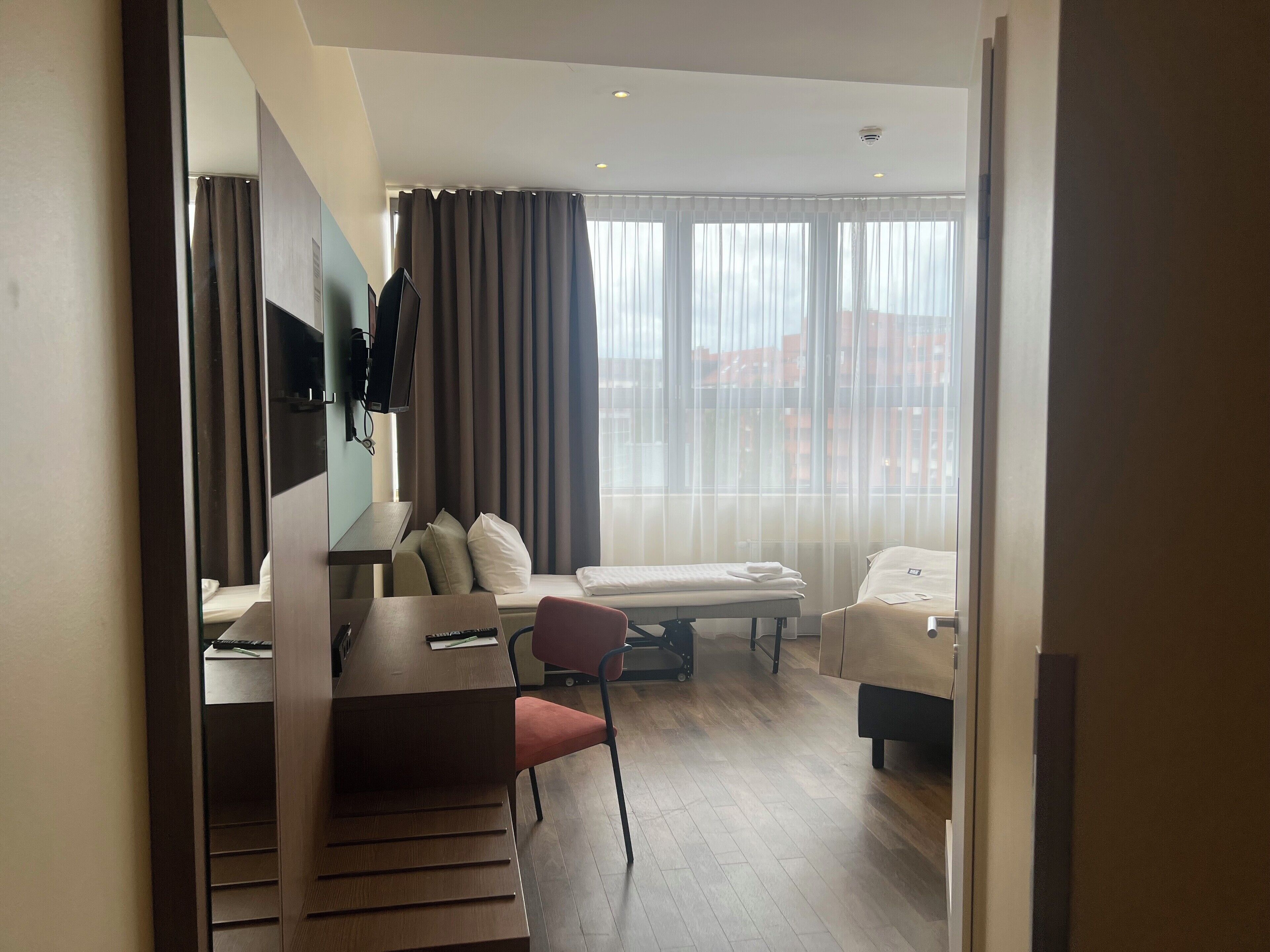 Una habitación de hotel moderna con escritorio, cama, silla y un amplio ventanal con cortinas.