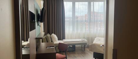 A modern hotel room with a desk, a bed, a chair, and a large window with curtains.
