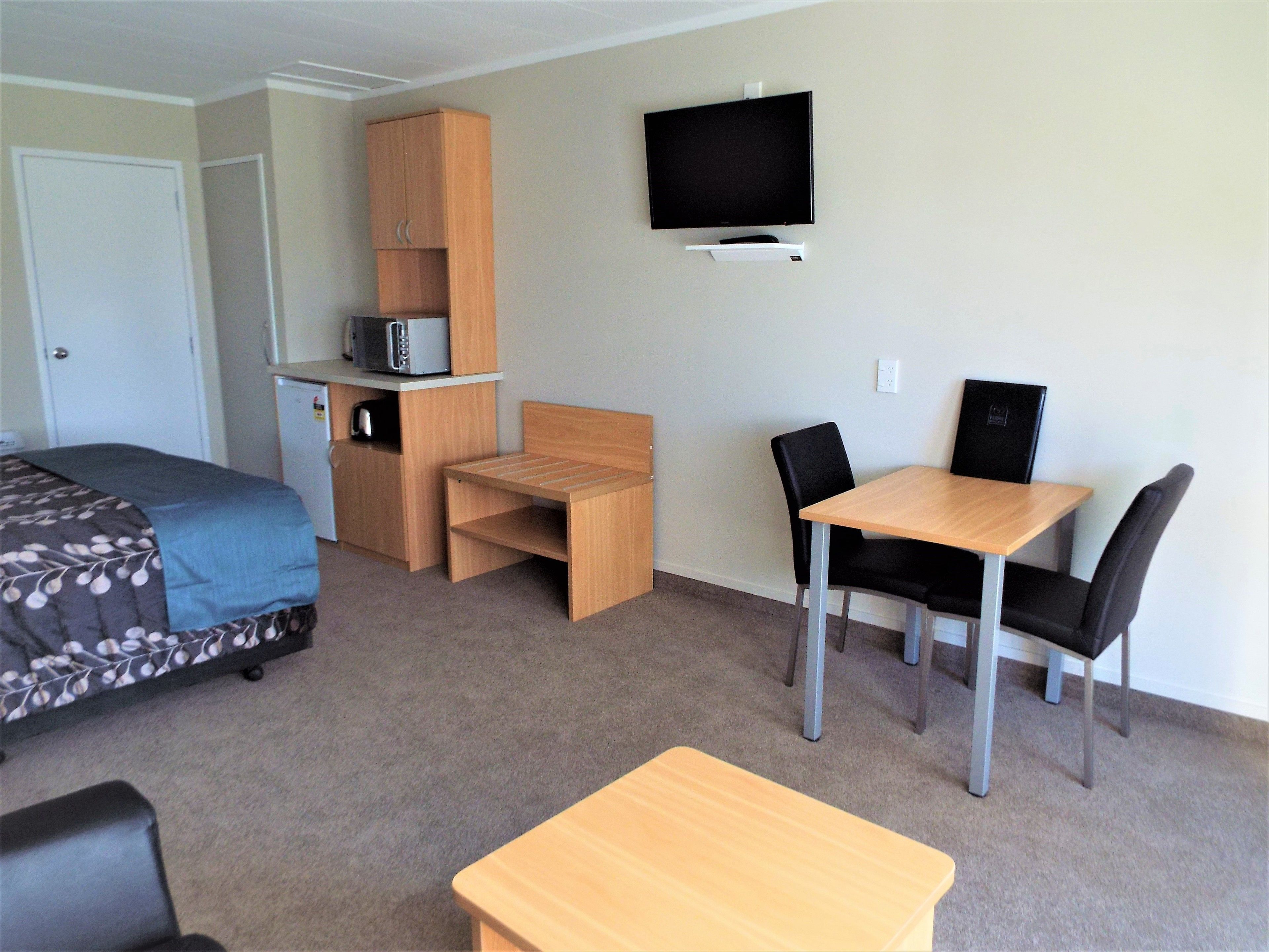 A compact hotel room with a bed, a small desk, a chair, a TV mounted on the wall, and a microwave on a shelf.