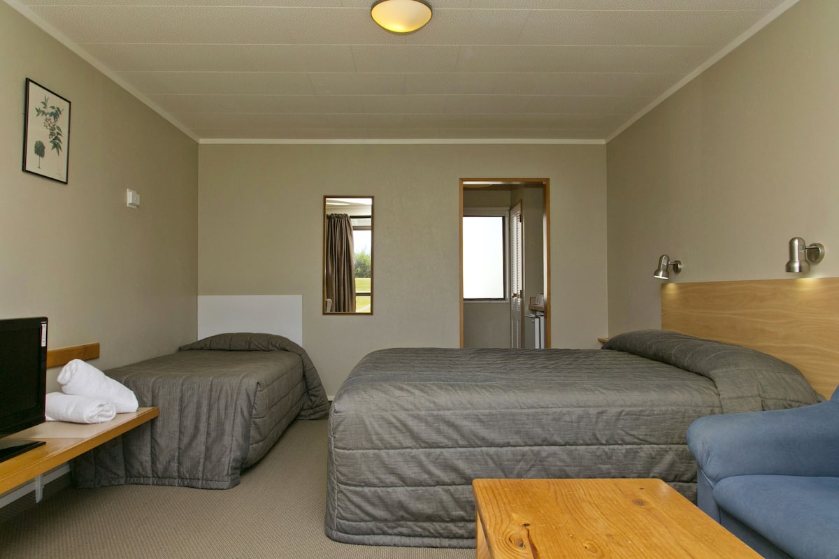 A hotel room with two single beds, a wooden desk with a television, a small table, and a blue chair.