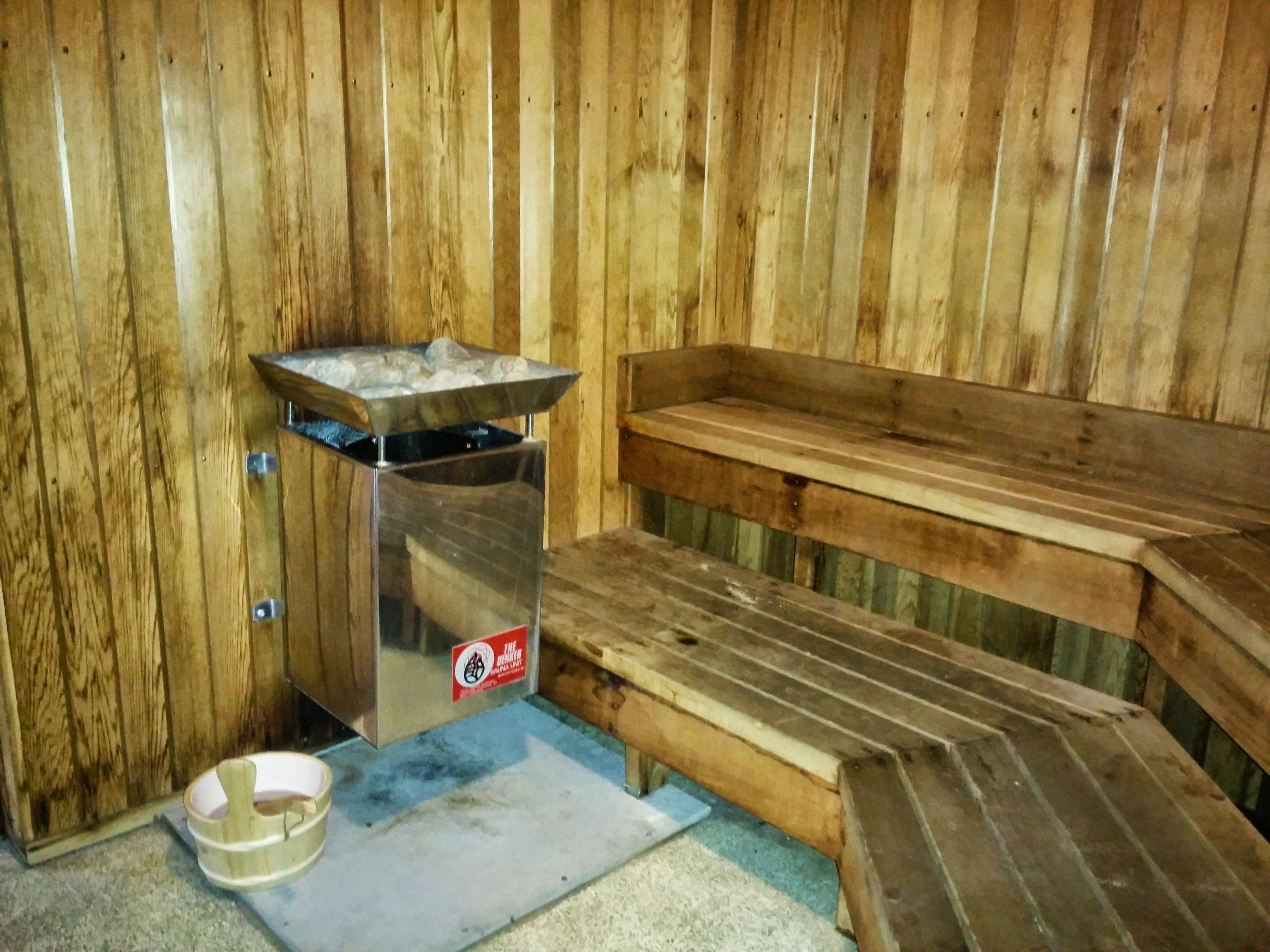 A sauna room with a wooden bench, a metal heater, and a small trash bin.