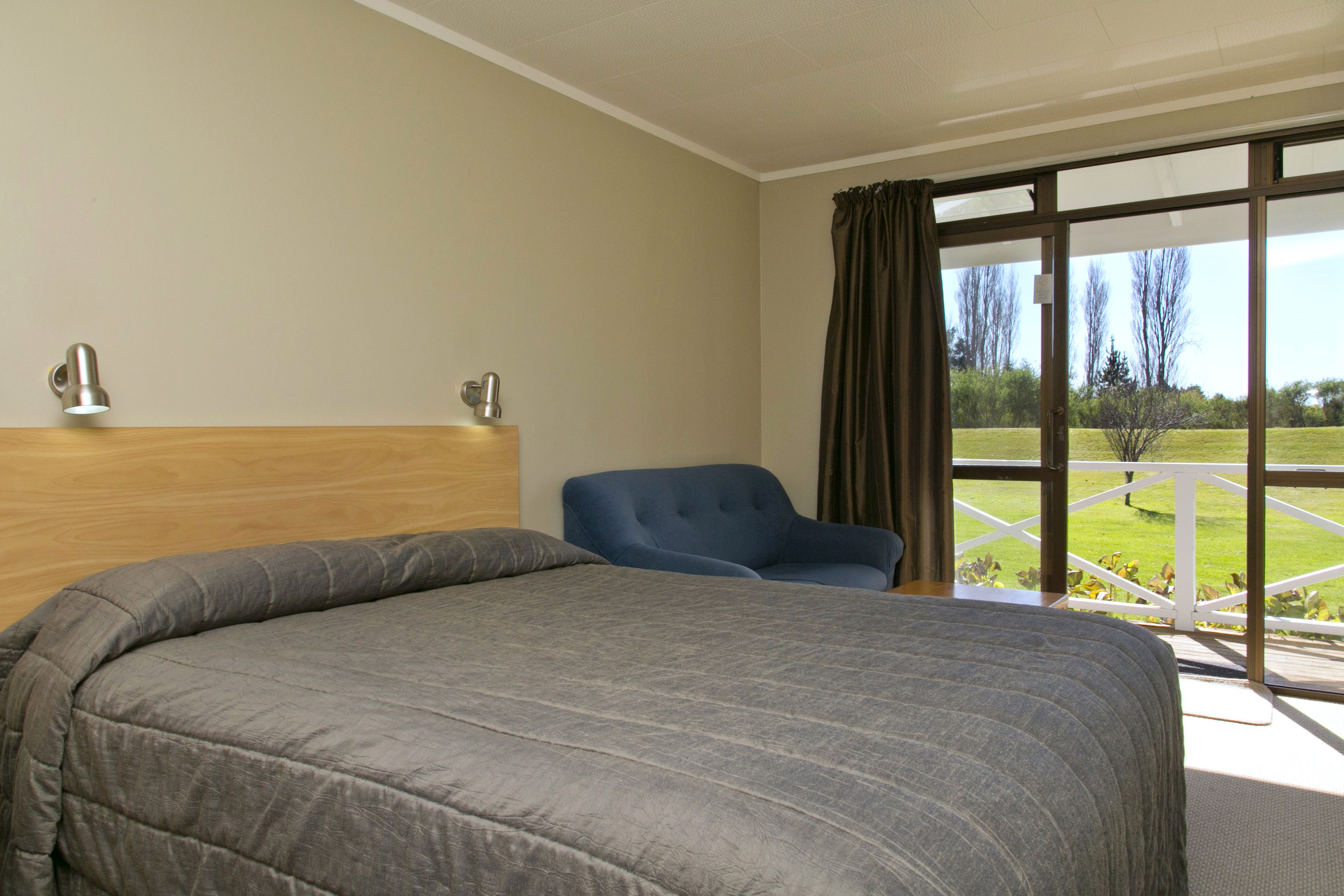 Habitación de hotel con una cama grande, una silla azul y vistas a un paisaje verde a través de un ventanal.
