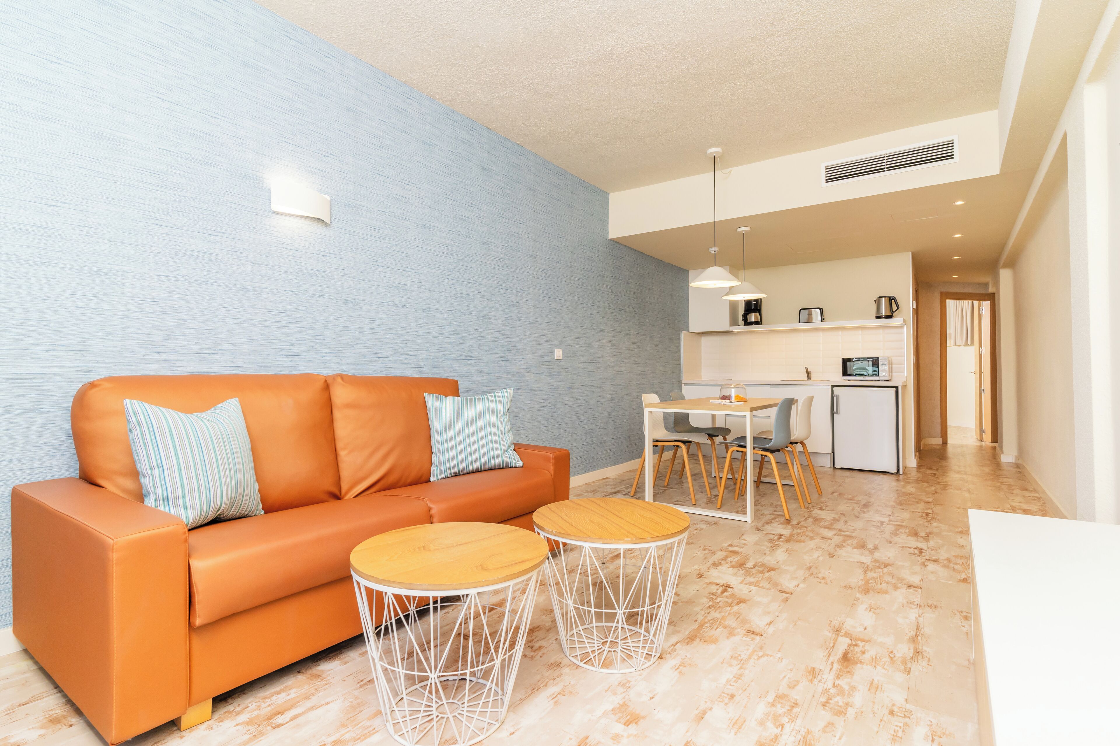 A modern living room with a sofa, two round tables, a kitchen area with a dining table and chairs, and a visible door leading to another room.