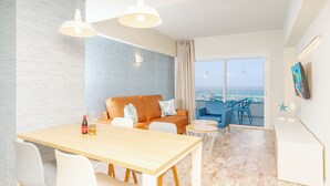 Una sala de estar moderna con una mesa de comedor de madera, sillas blancas, un sofá con cojines naranja y vistas al mar a través de ventanas amplias.