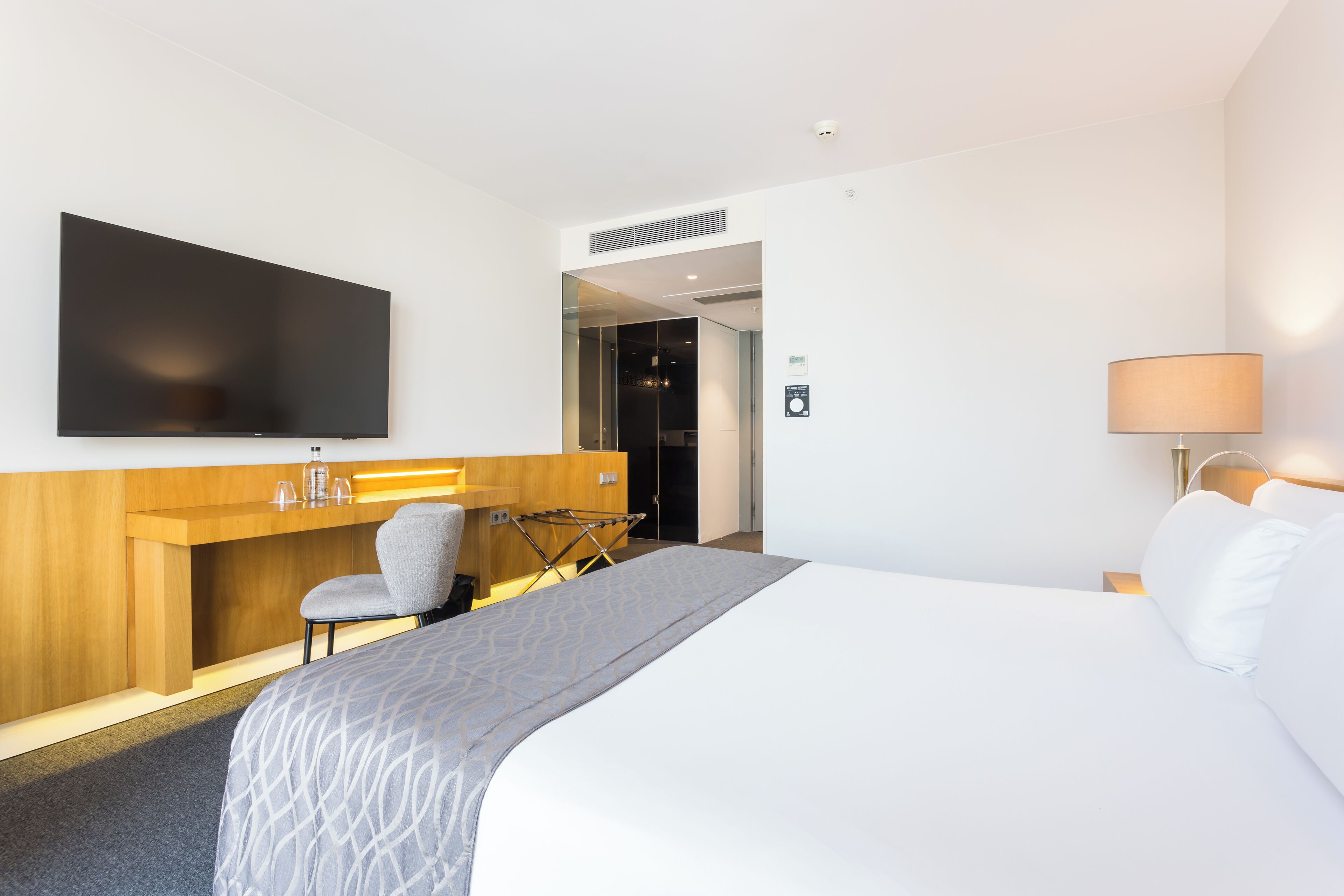 A hotel room with a large bed, a desk with a flat-screen TV, a chair, a lamp, and a wardrobe.