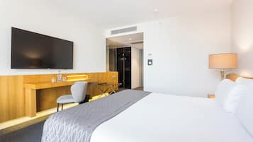 A hotel room with a large bed, a desk with a flat-screen TV, a chair, a lamp, and a wardrobe.