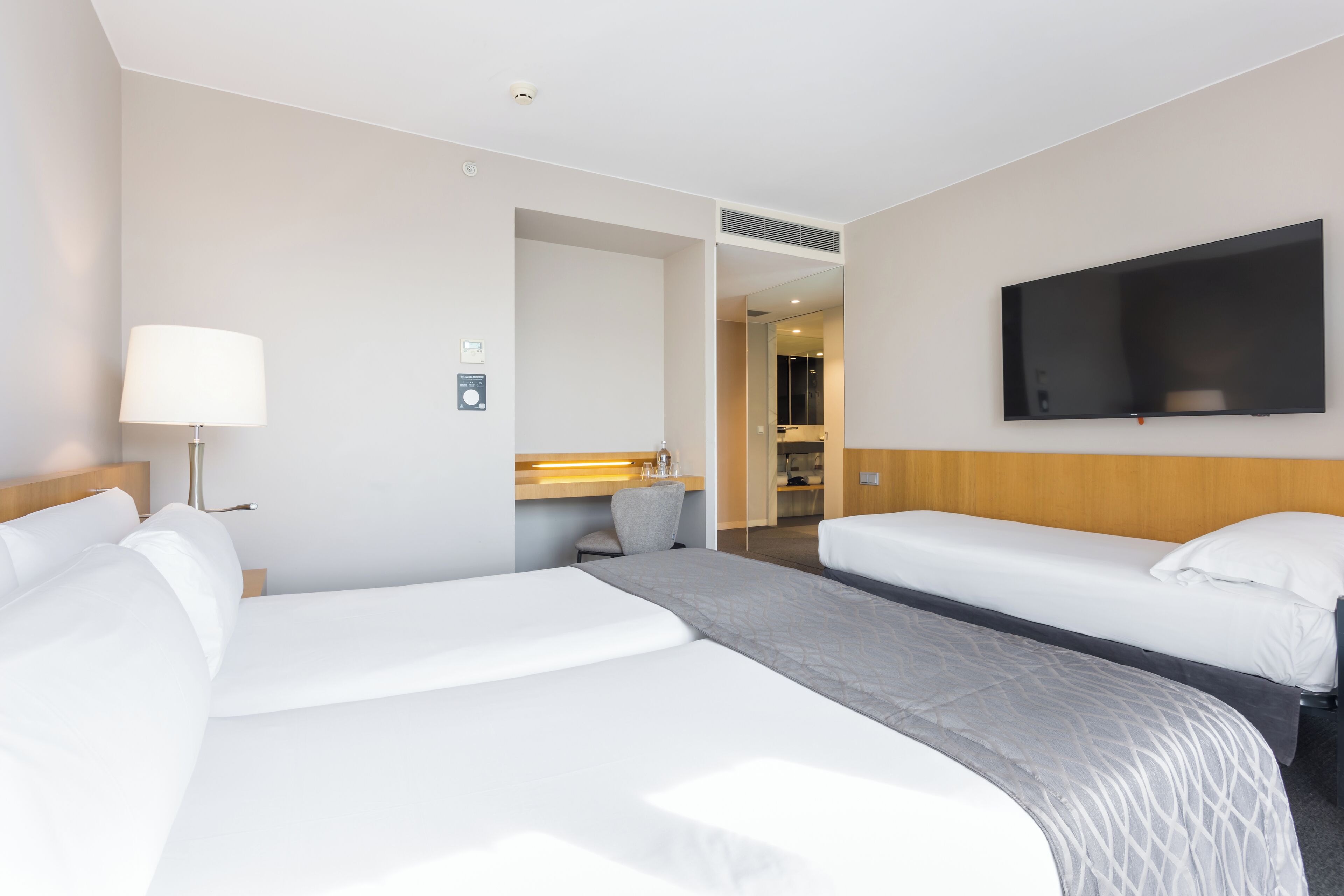 A hotel room with two beds, a TV mounted on the wall, a bedside table with a lamp, and a bathroom visible through an open door.