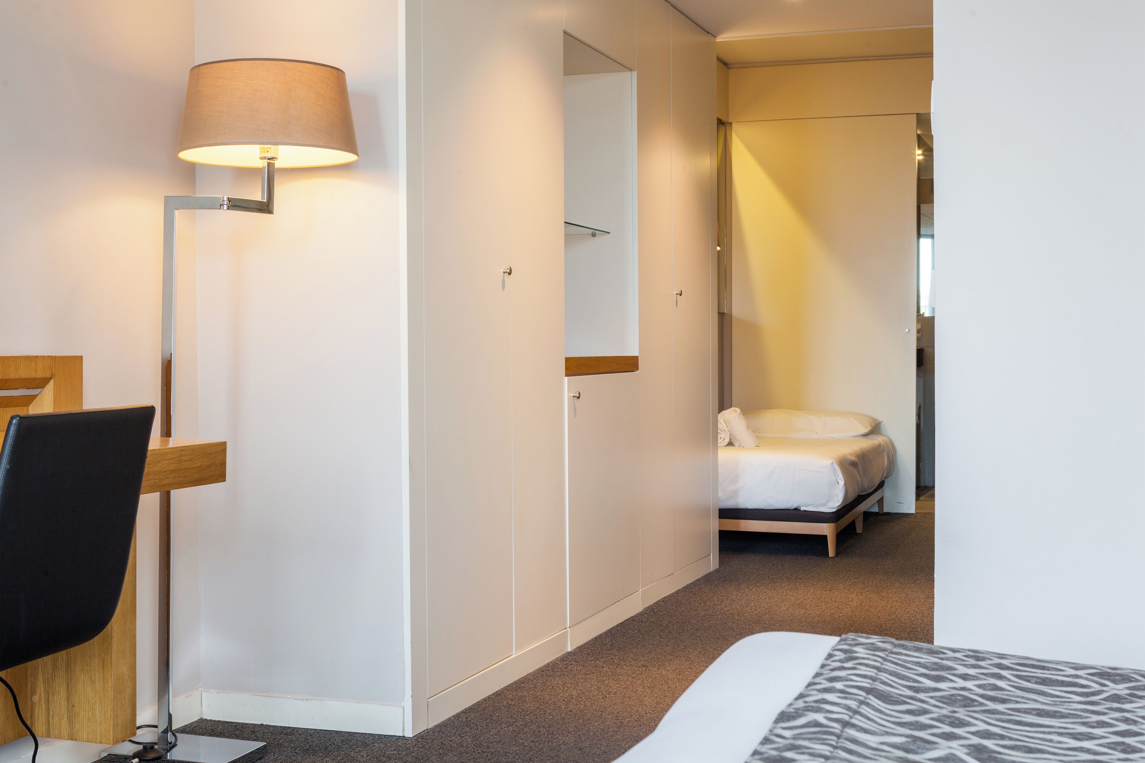 A hotel room with a bed, a desk with a chair, a lamp, a wardrobe, and a mirror.
