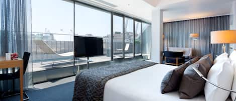 Una habitación de hotel con una cama grande, un escritorio, una silla, un televisor y vistas a la ciudad a través de amplias ventanas.