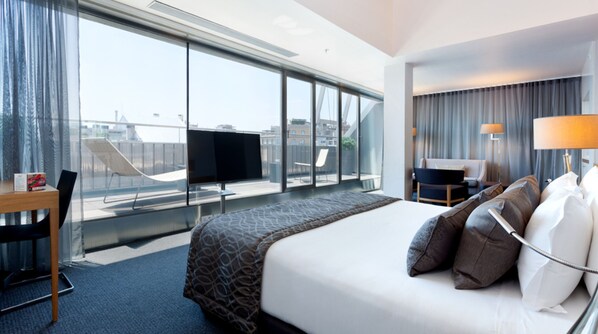 Una habitación de hotel con una cama grande, un escritorio, una silla, un televisor y vistas a la ciudad a través de amplias ventanas.