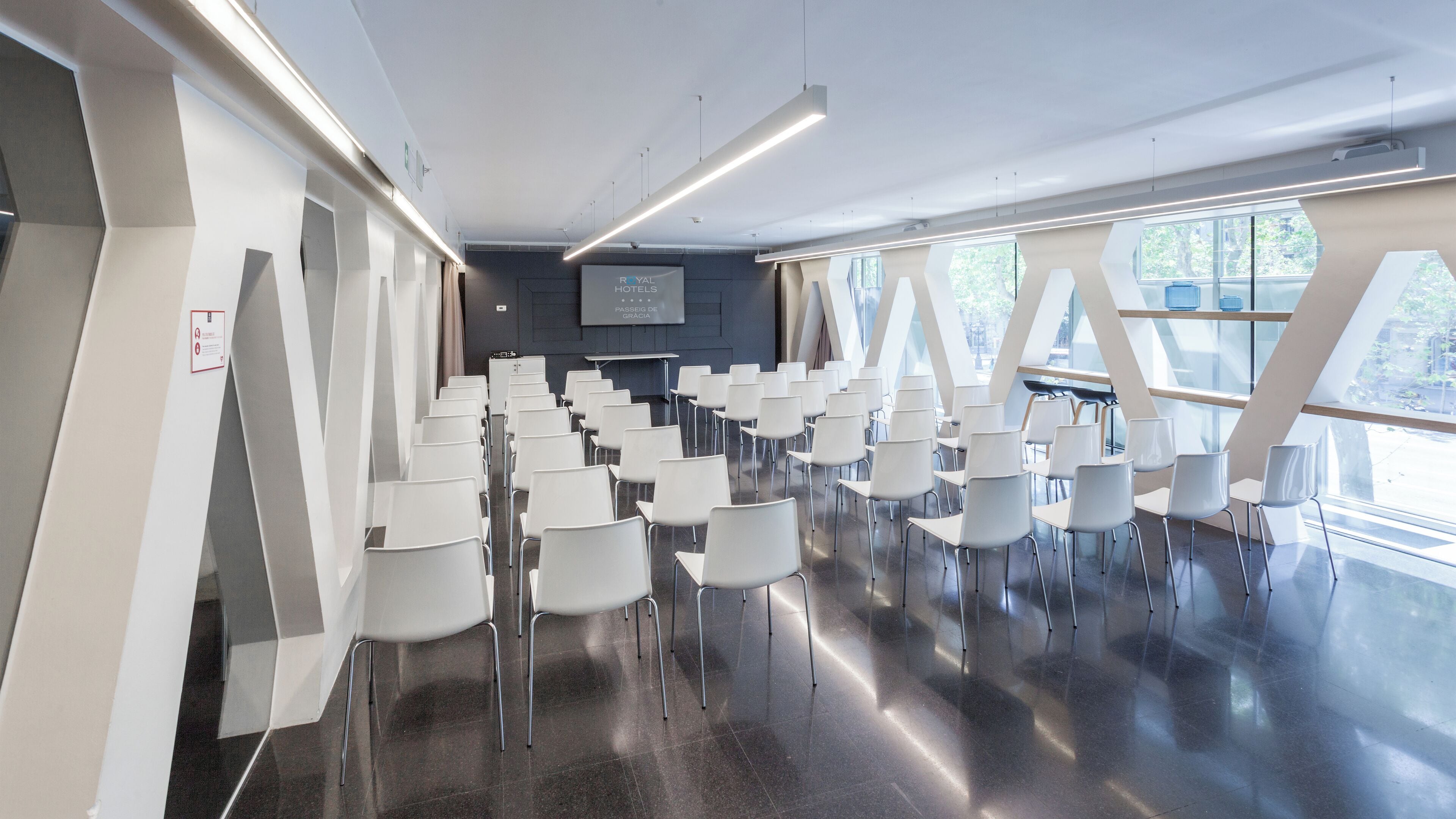 Une salle de conférence moderne, dotée de rangées de chaises blanches, d’un grand écran et d’un mur de verre offrant une vue sur l’extérieur.
