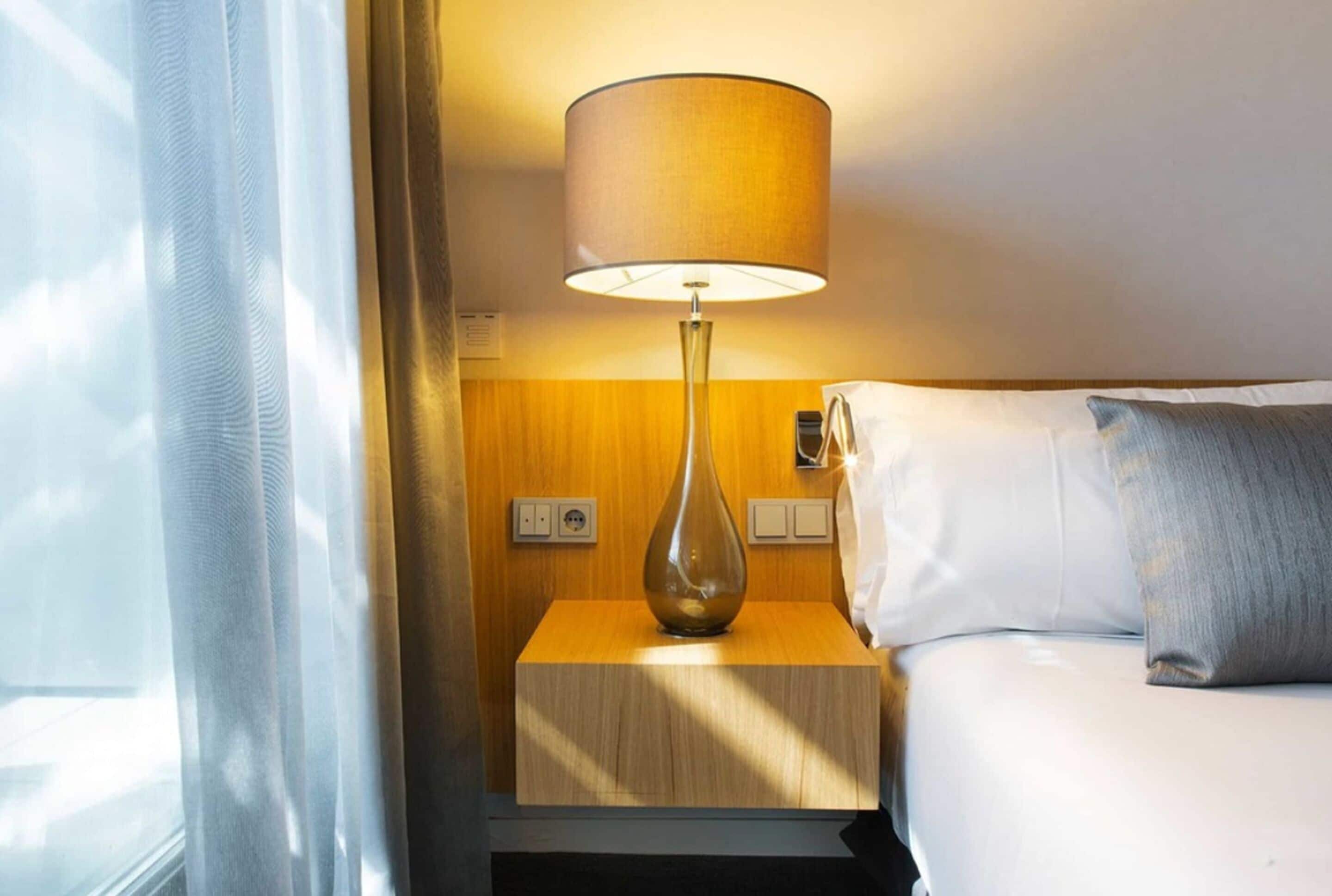 Una habitación de hotel con una cama, una lámpara de noche, una mesita de noche y una ventana con cortinas.