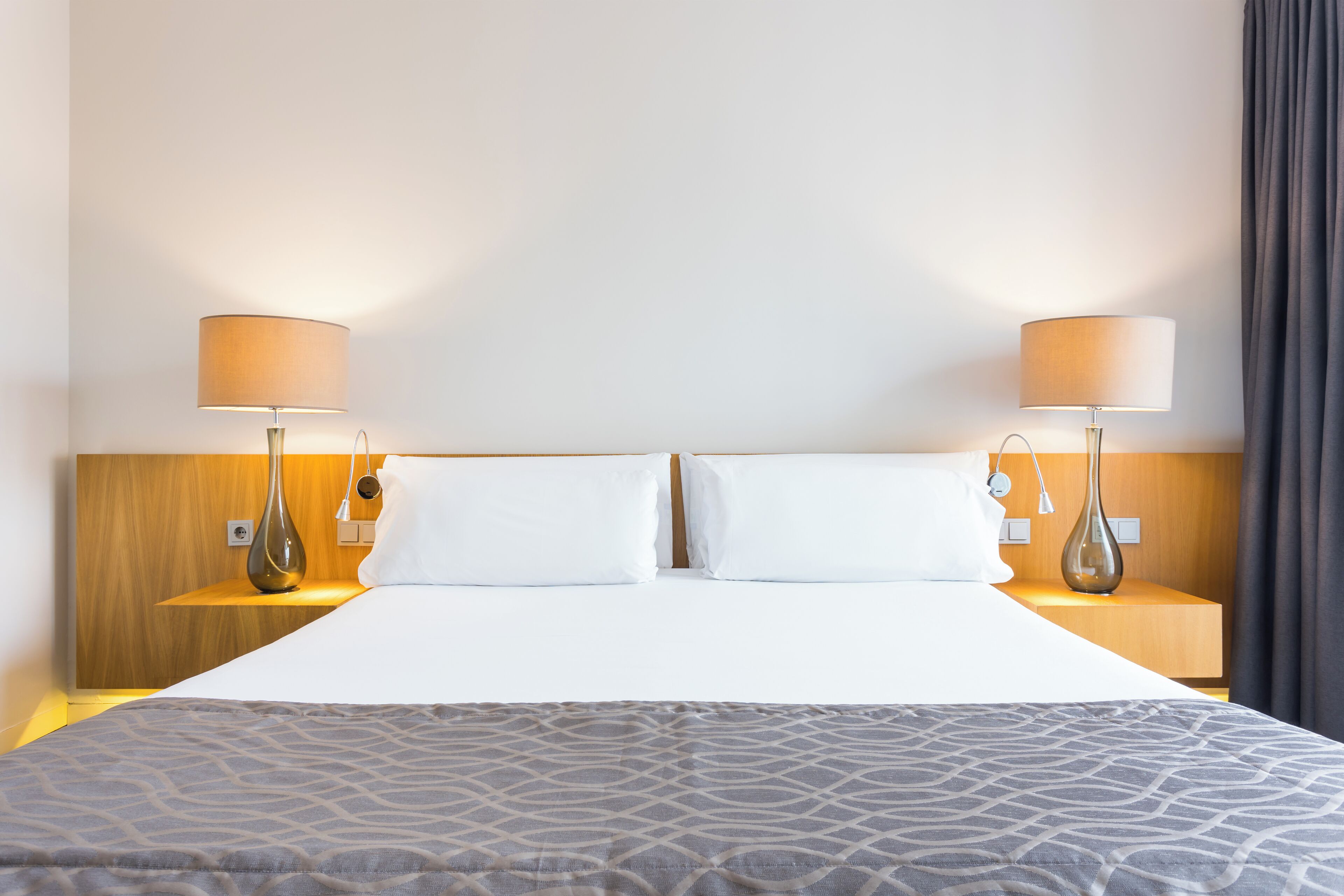Une chambre d’hôtel avec un grand lit, deux lampes de chevet, une tête de lit en bois et une couvre-lit grise à motifs.
