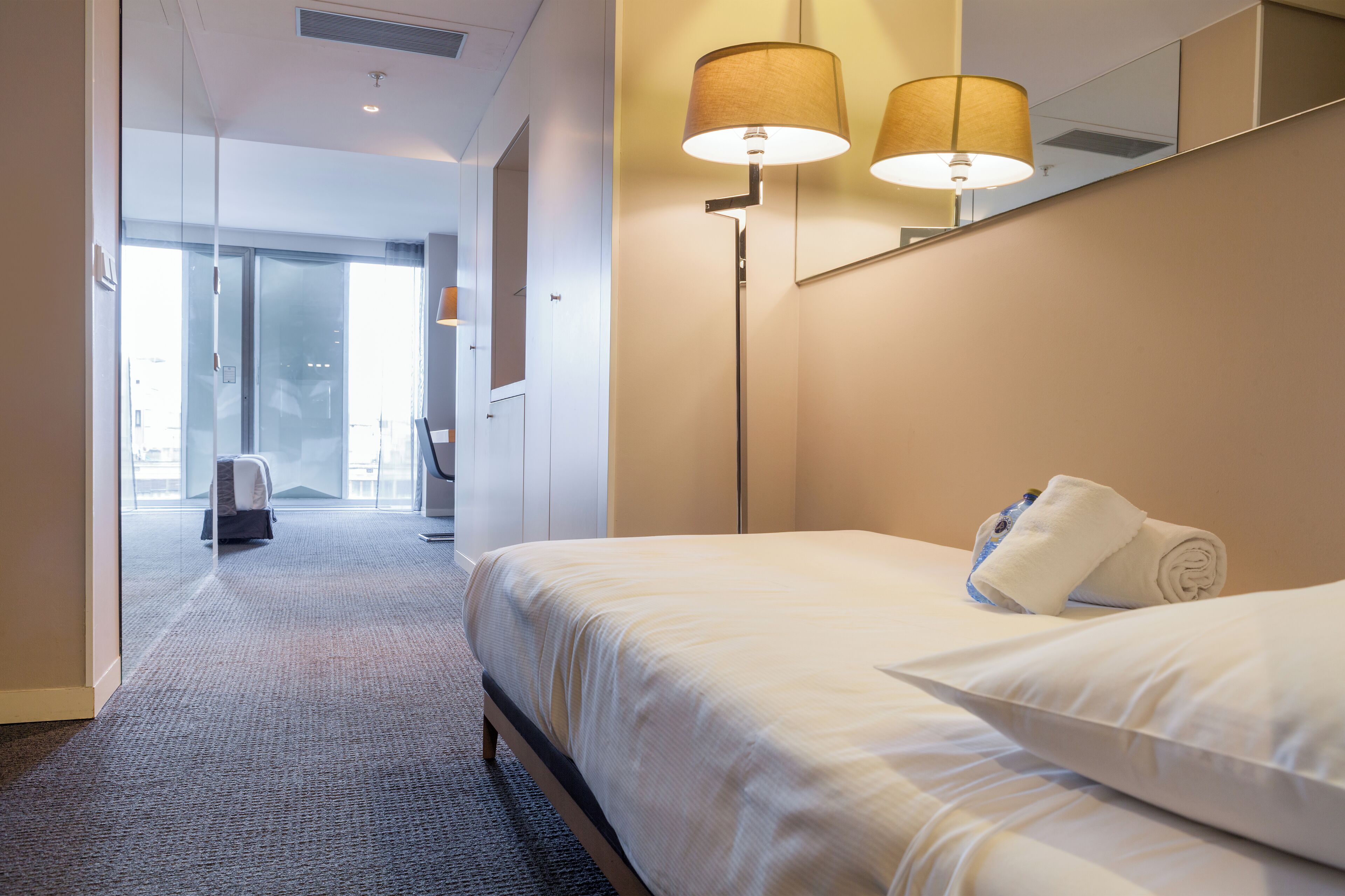 Una habitación de hotel con una cama bien hecha, un espejo grande y una lámpara de pie.