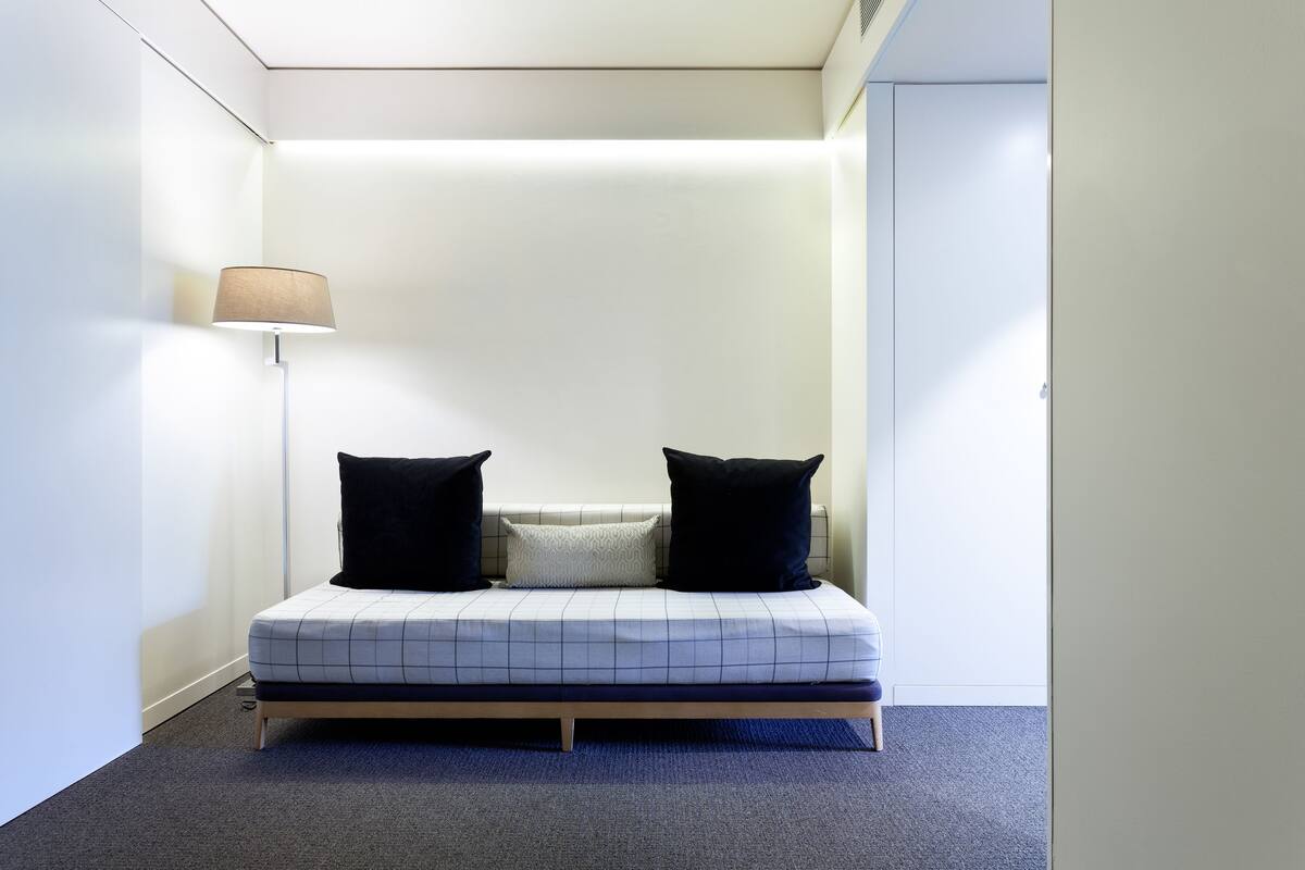 A small, neatly arranged bedroom with a single bed, a bedside lamp, and a plain wall.