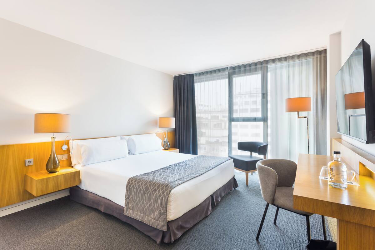 A hotel room with a bed, bedside tables, a desk, a chair, and a TV.