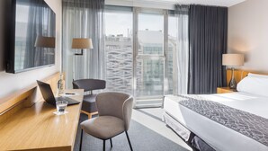 Habitación de hotel con cama, escritorio con laptop, silla, televisor y ventana grande con cortinas.