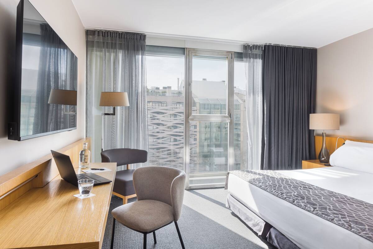 A hotel room with a bed, a desk with a laptop, a chair, a TV, and a large window with curtains.