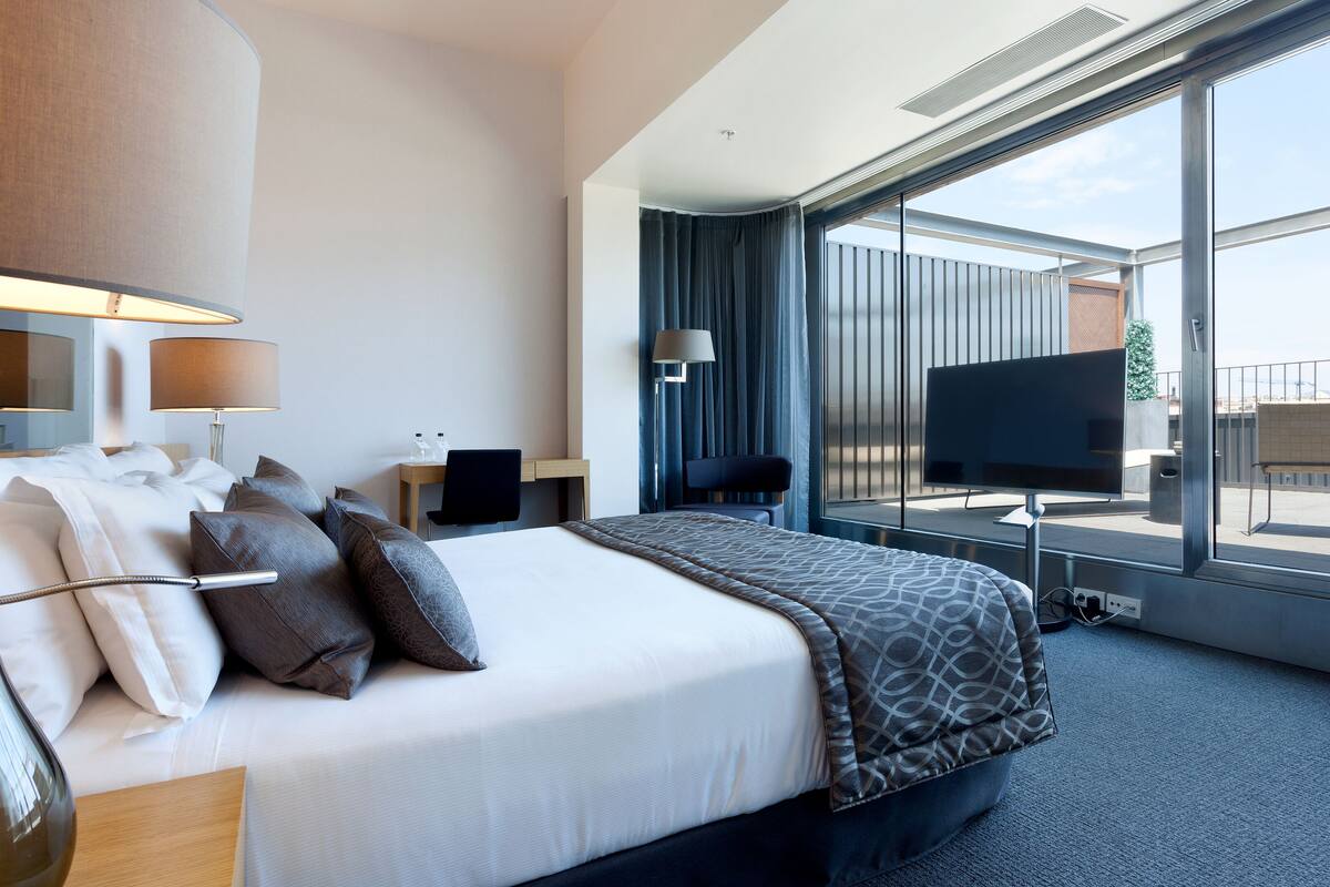 A modern hotel room with a large bed, a desk, a chair, a TV, and a balcony with a view.
