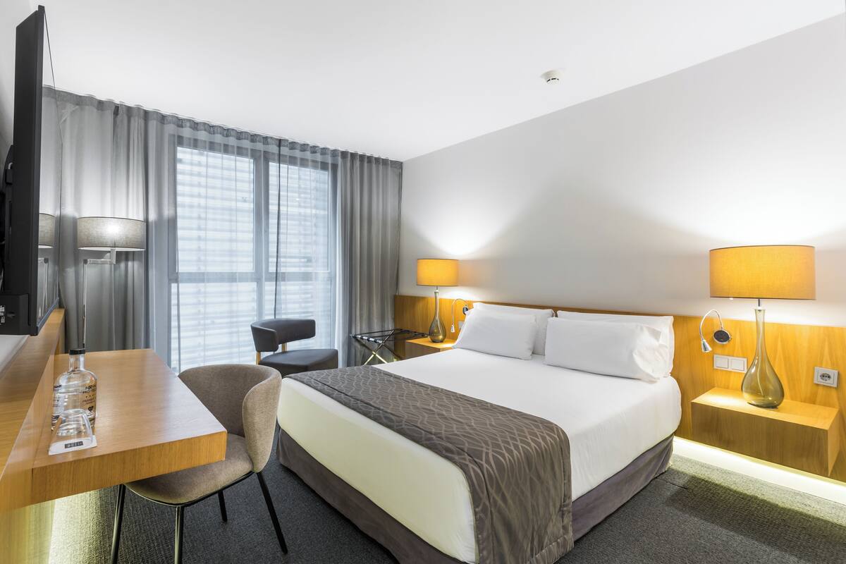 A hotel room with a bed, a desk, a chair, a TV, and two bedside lamps.