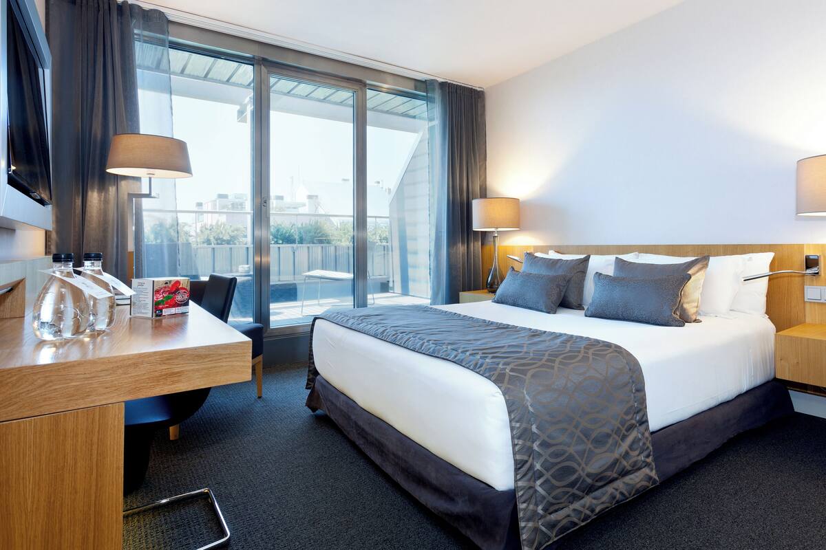 A hotel room with a large bed, two bedside lamps, a desk with a chair, a small table with water bottles, and a view of the city through large windows.