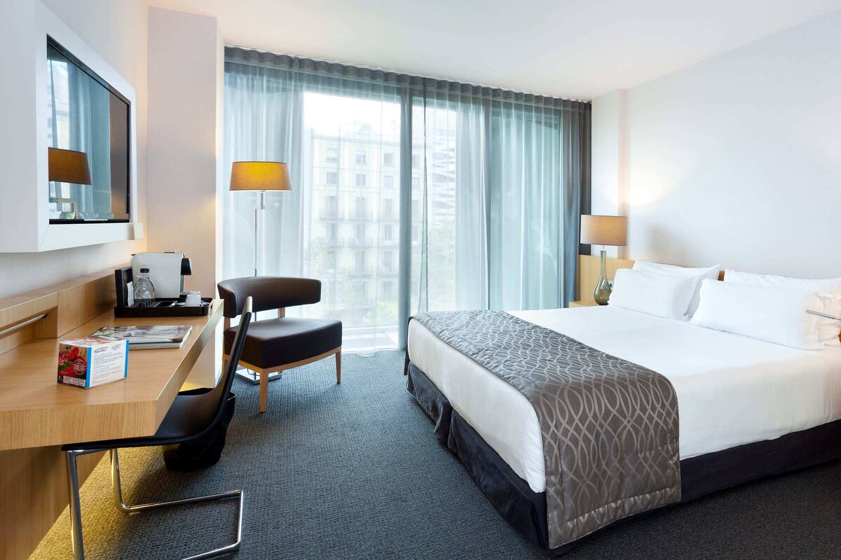 A hotel room with a large bed, two bedside tables with lamps, a desk with a chair, a TV, and a window with curtains.