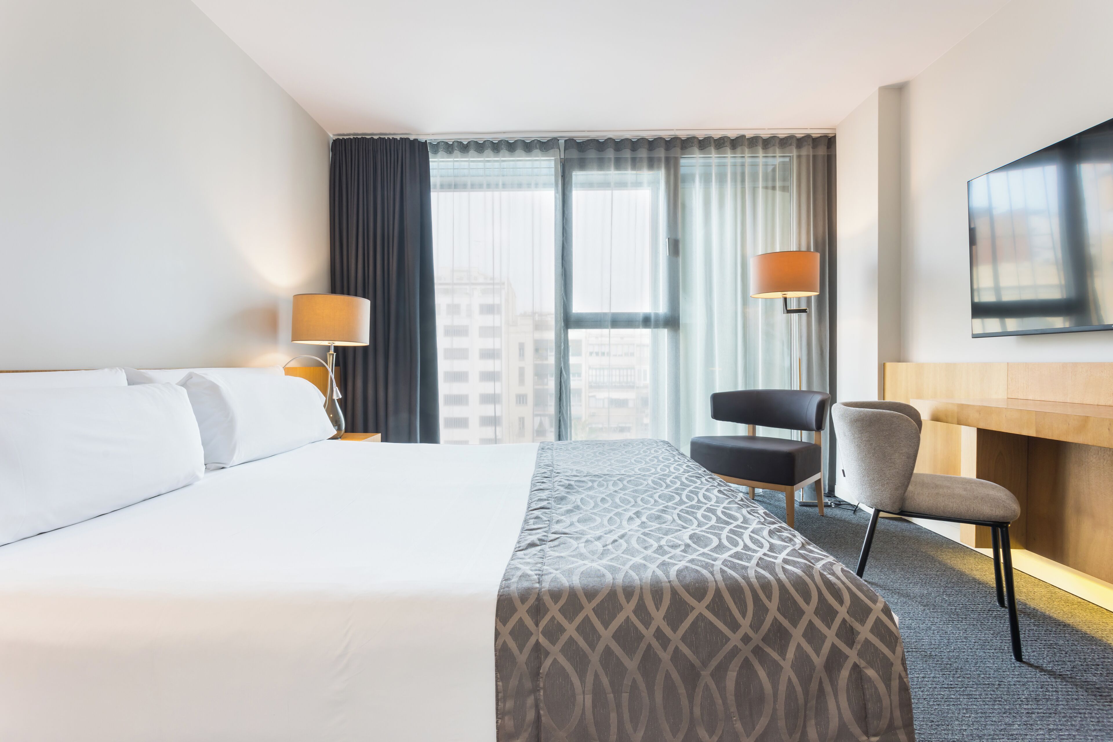 A hotel room with a large bed, two bedside lamps, a desk with a chair, and a large window with curtains.