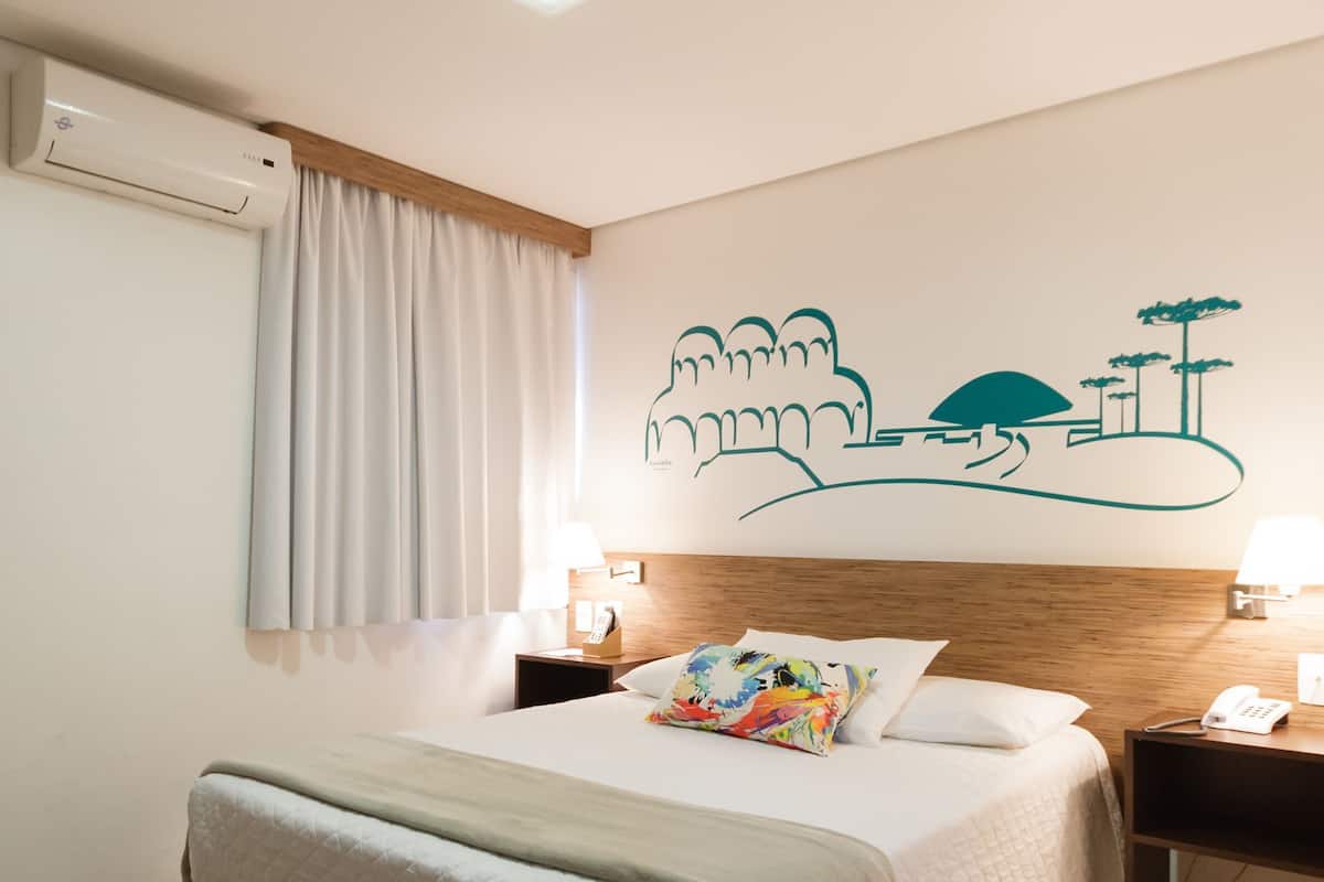 A hotel room with a bed, a nightstand, a wall-mounted air conditioner, and a window with curtains.
