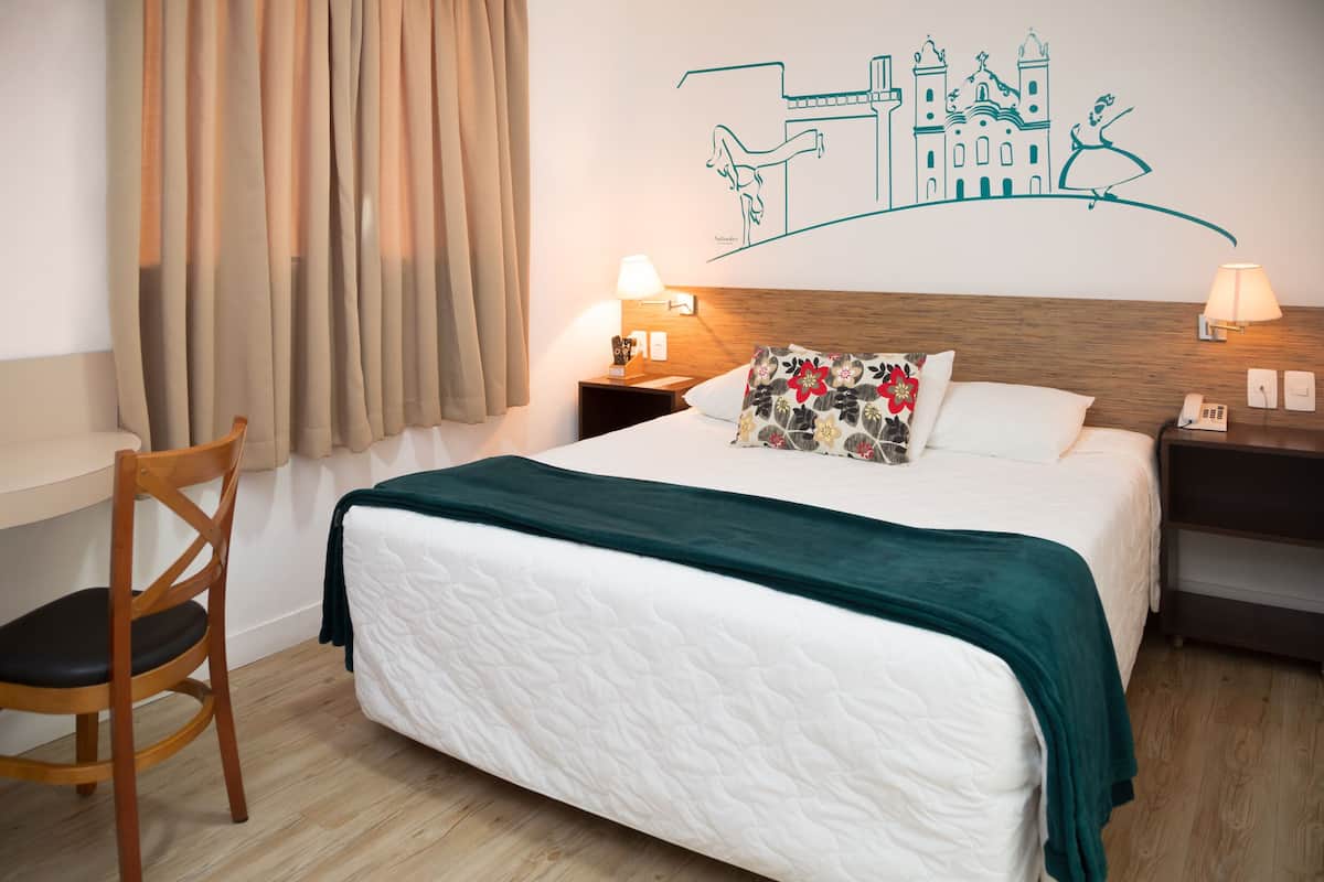 A hotel room with a bed, a wooden nightstand, a chair, and a wall mural.