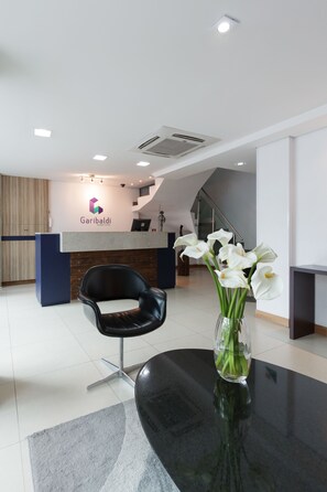 A reception area with a modern desk, a black chair, and a vase with white flowers. - Garibaldi Business Hotel (Porto Alegre)