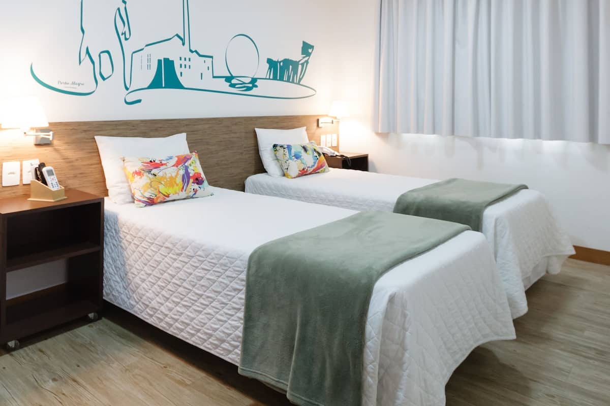 A hotel room with two beds, a cityscape wall art, a wooden floor, and a window with curtains.