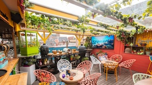 An outdoor seating area with wooden tables and colorful chairs, a green counter with fruit, and a TV screen displaying an underwater scene. - The Happy Traveler (Groningen)
