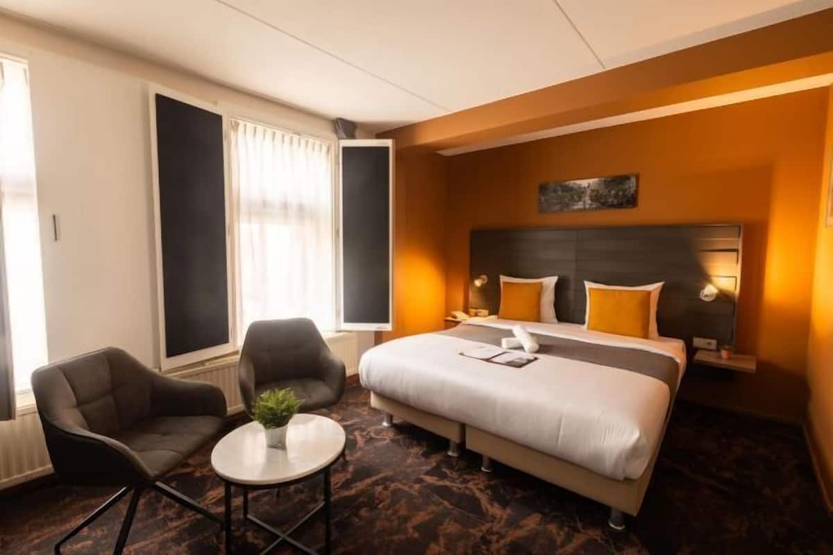 A hotel room with a bed, two armchairs, a small table with a plant, a window with curtains, and a headboard with a light fixture.