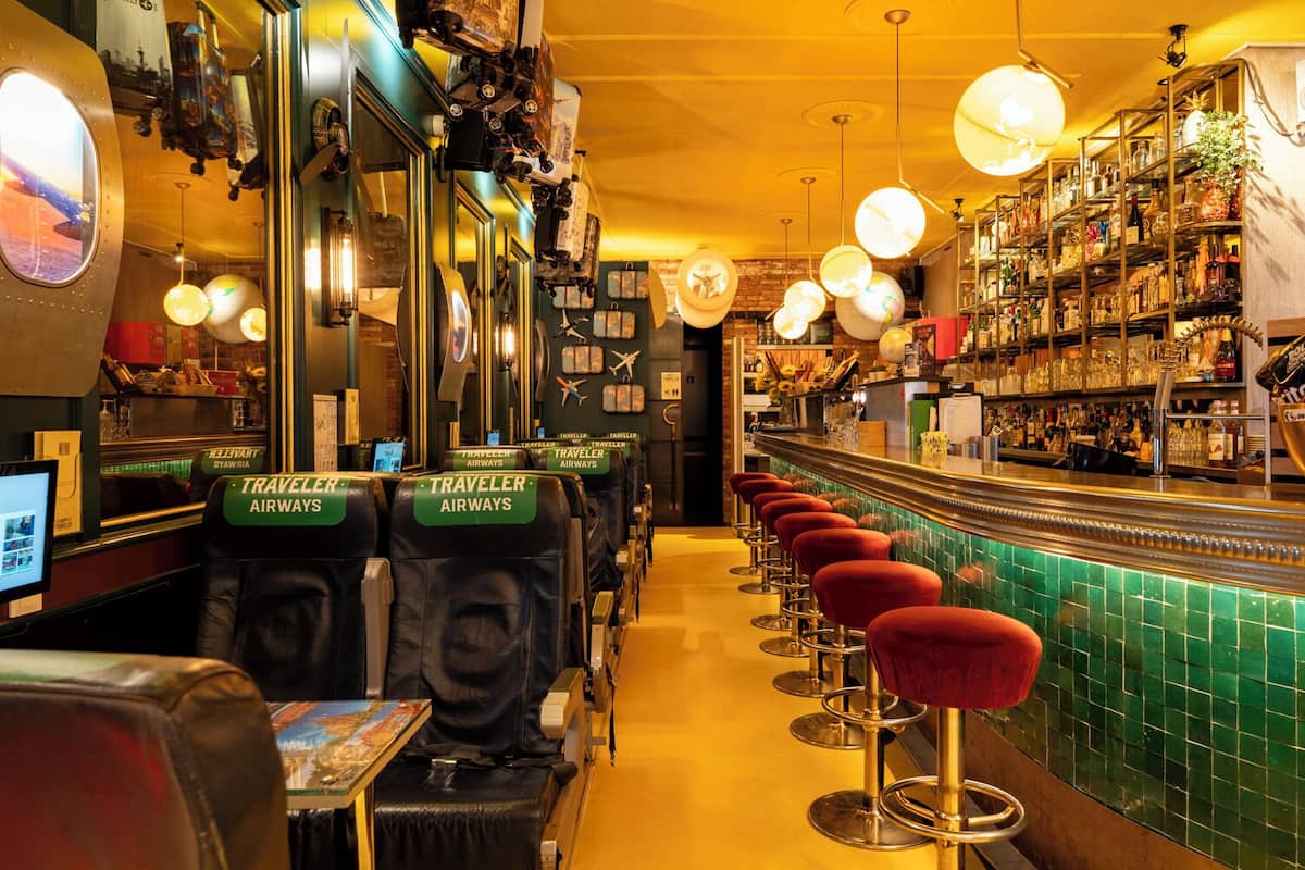 A bar area with a green-tiled counter, red bar stools, and a row of seats labeled 'TRAVELER AIRWAYS'.