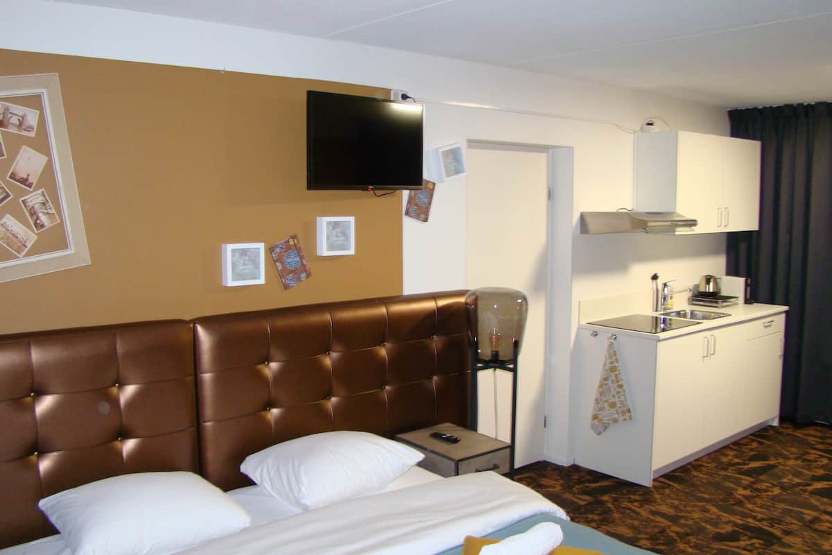 A hotel room with a brown tufted headboard bed, a flat-screen TV mounted on the wall, a small bedside table with a remote control, a kitchenette with a sink and cabinets, and a patterned carpet.