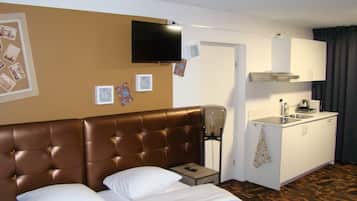 A hotel room with a brown tufted headboard bed, a flat-screen TV mounted on the wall, a small bedside table with a remote control, a kitchenette with a sink and cabinets, and a patterned carpet.
