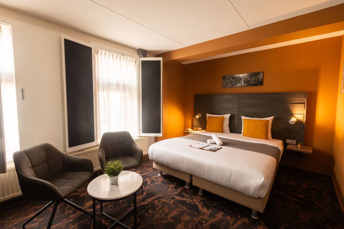 A hotel room with a bed, two armchairs, a small table with a plant, and a window with curtains.