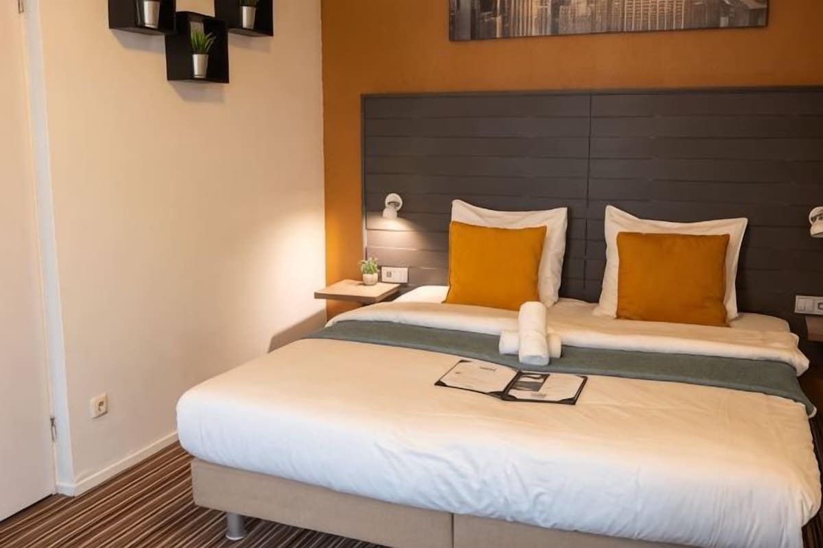 A hotel room with a large bed, a nightstand, a wall-mounted lamp, and a small potted plant.