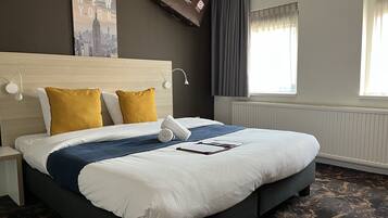 A hotel room with a large bed, a headboard, a wall-mounted TV, a window with curtains, and a radiator.
