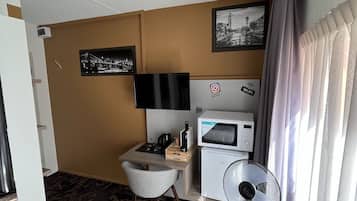 A compact hotel room with a bed, a desk with a chair, a small refrigerator, a microwave, a fan, and a picture on the wall.