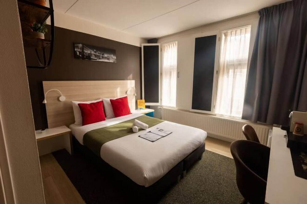 A hotel room with a bed, a nightstand, a desk, a chair, and a window with curtains.