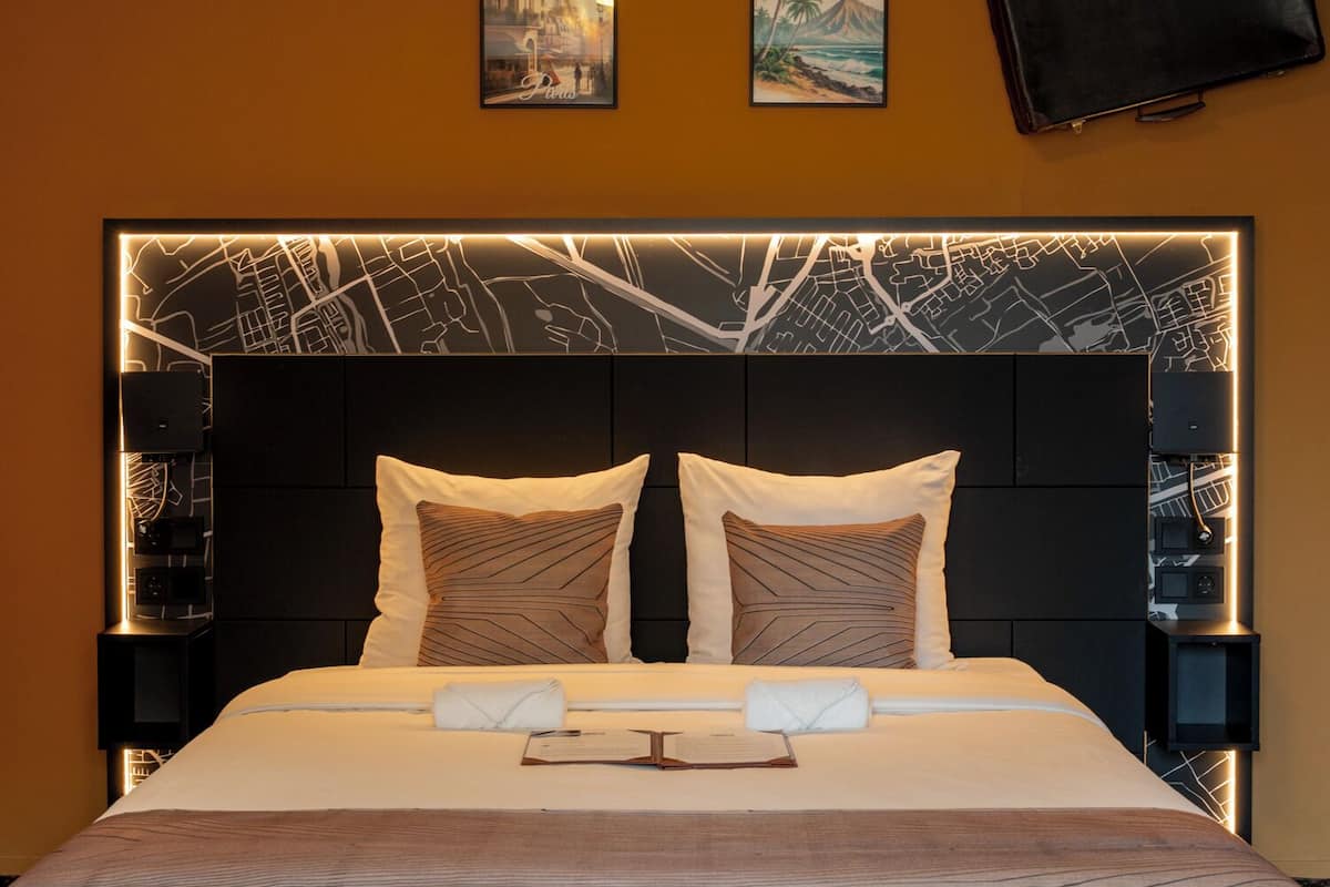 A hotel room with a large bed, two bedside tables, a headboard with a cityscape design, and a flat-screen TV mounted on the wall.