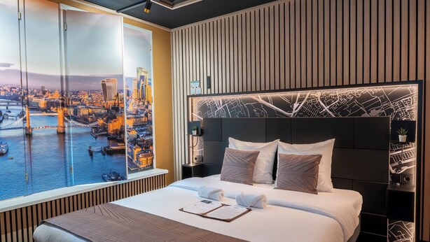 A hotel room with a large window, a bed with pillows, a nightstand, and a wall with a map design. - The Happy Traveler (Groningen)