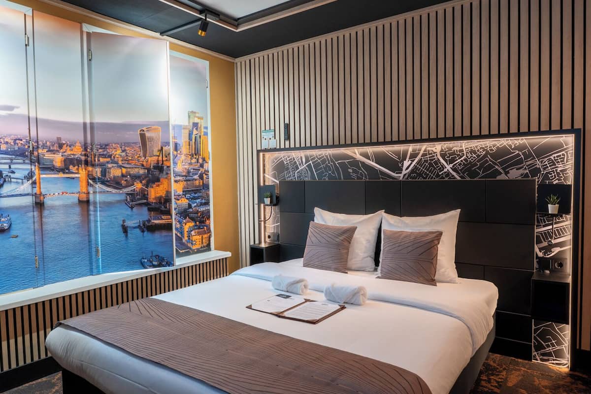 A hotel room with a large window, a bed with pillows, a nightstand, and a wall with a map design.