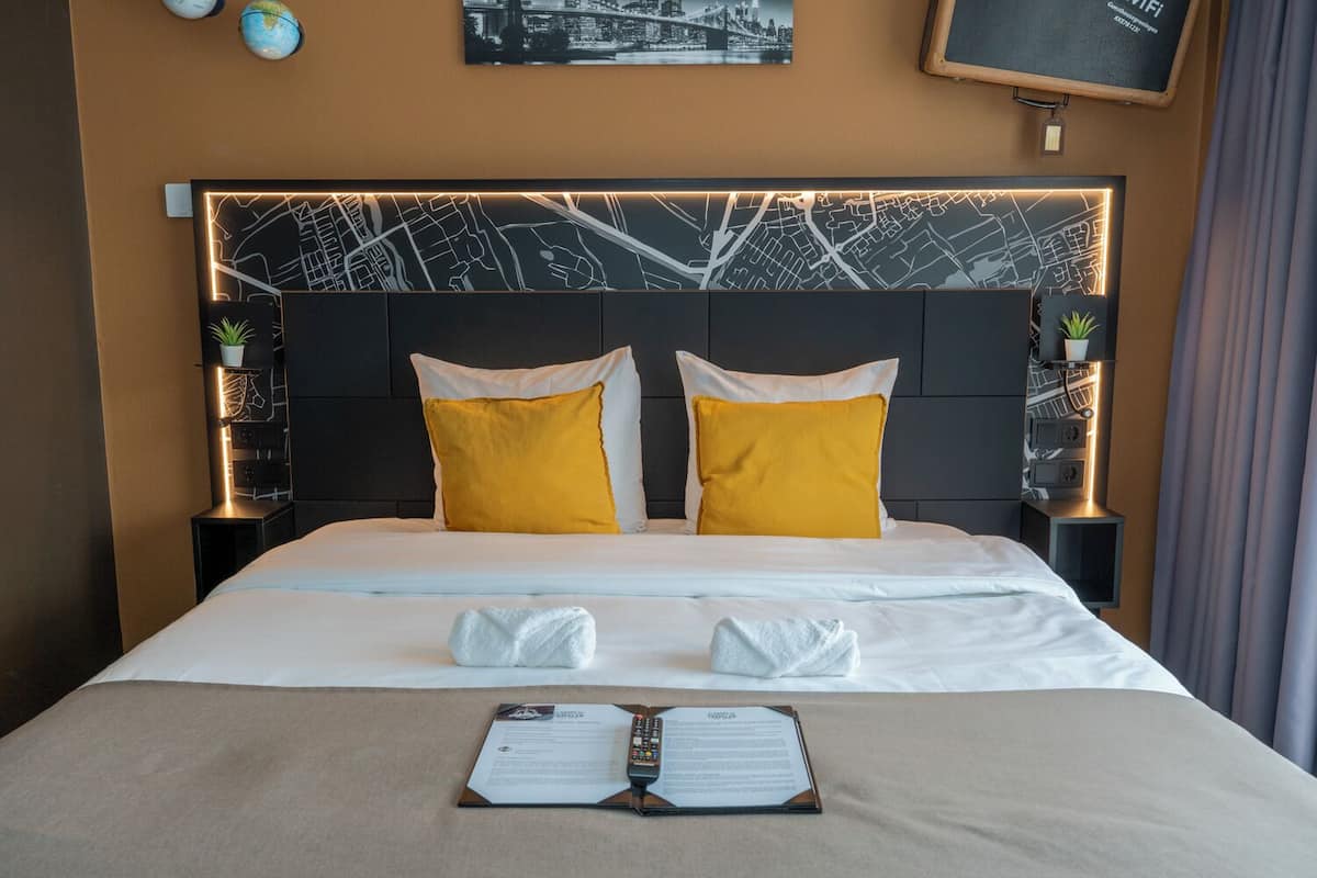 A hotel room with a large bed, two pillows, a bedside table with a lamp, a map on the wall, and a plant on the nightstand.