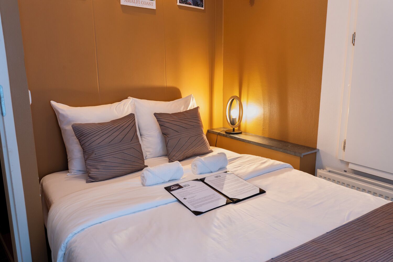 A hotel room with a bed, pillows, a nightstand, a mirror, and a door.